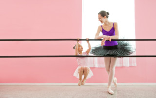 Little girl swings from ballet barre dressed in her ballerina outfit. Older ballerina in pointe shoes and platter tutus looks at her. Goal setting - how do we get form point A to pointe B?