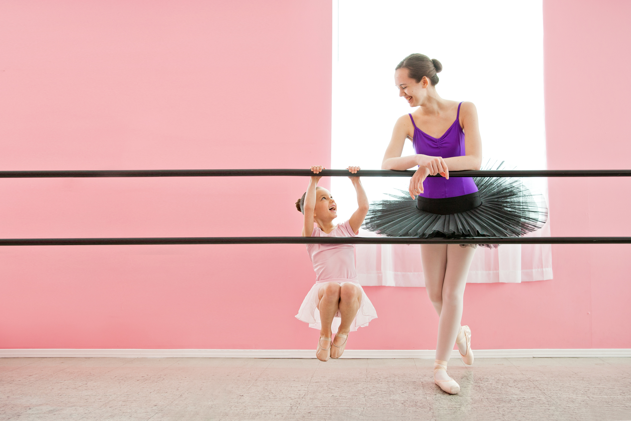 Little girl swings from ballet barre dressed in her ballerina outfit. Older ballerina in pointe shoes and platter tutus looks at her. Goal setting - how do we get form point A to pointe B?